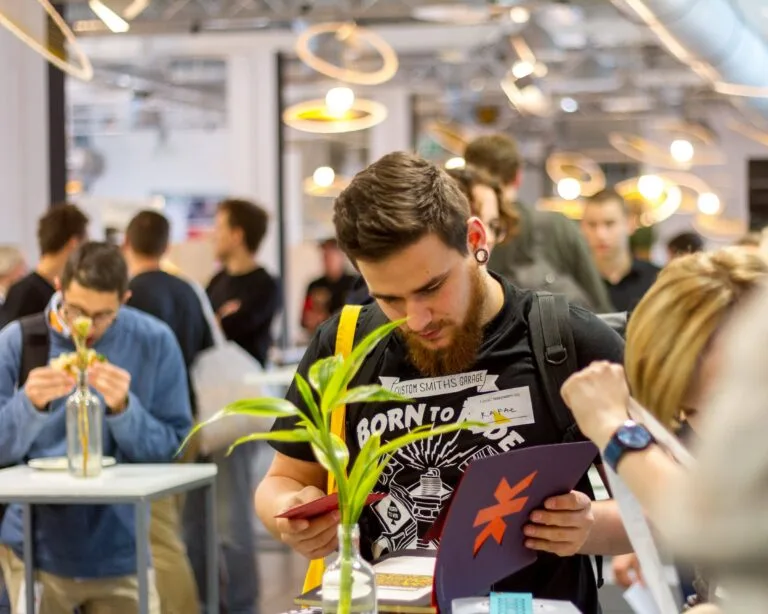 Man in a graphic jacket holding a plant and programme at a busy indoor event
