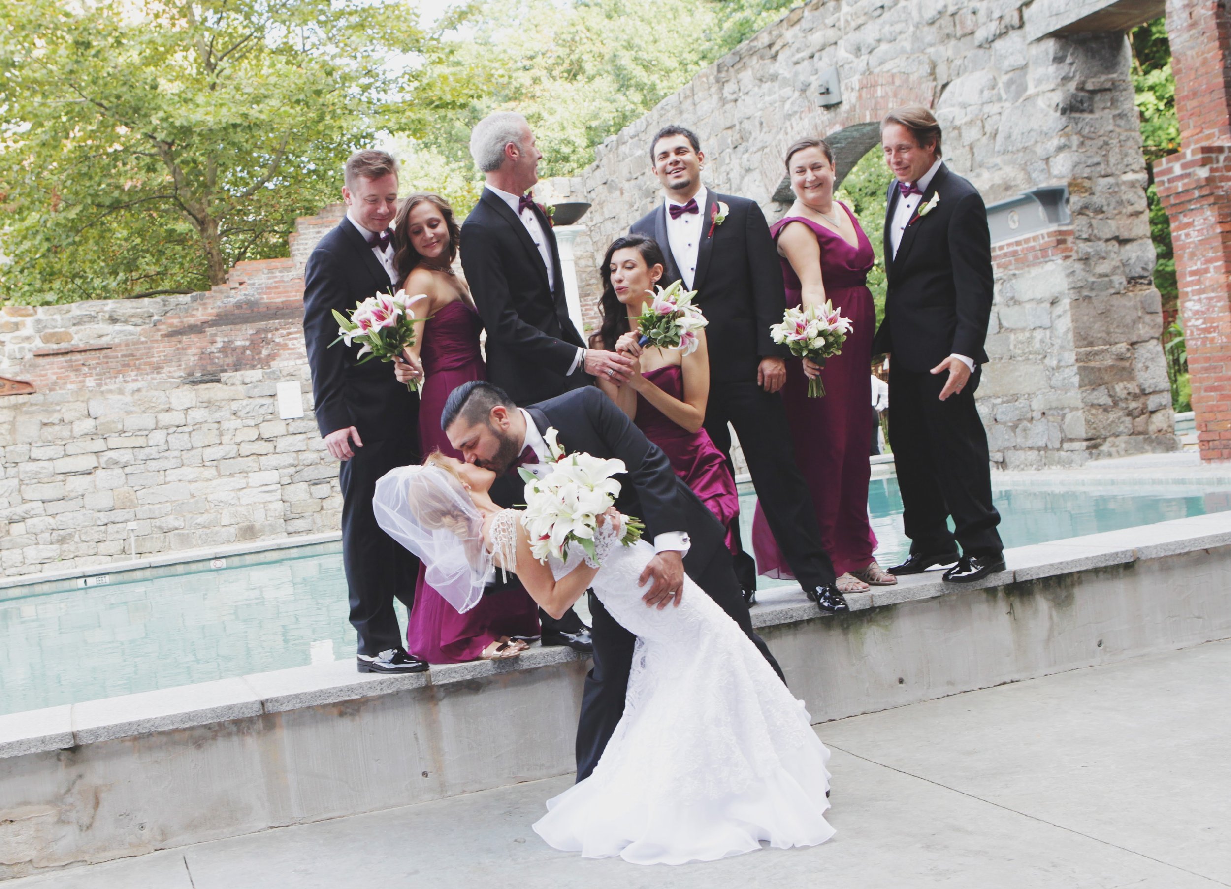 Groom dipping the bride for a kiss with the wedding party in burgundy behind them beside a pool