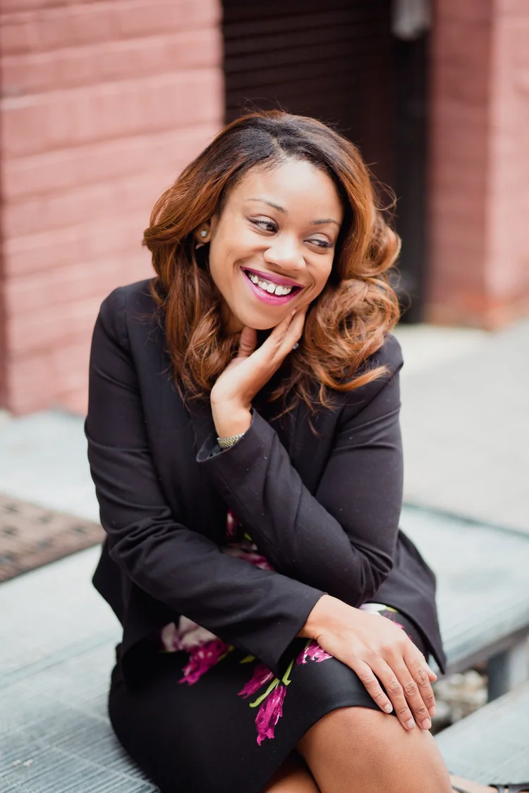 Andrea Davis smiling with her hand to her cheek, seated in front of a brick wall