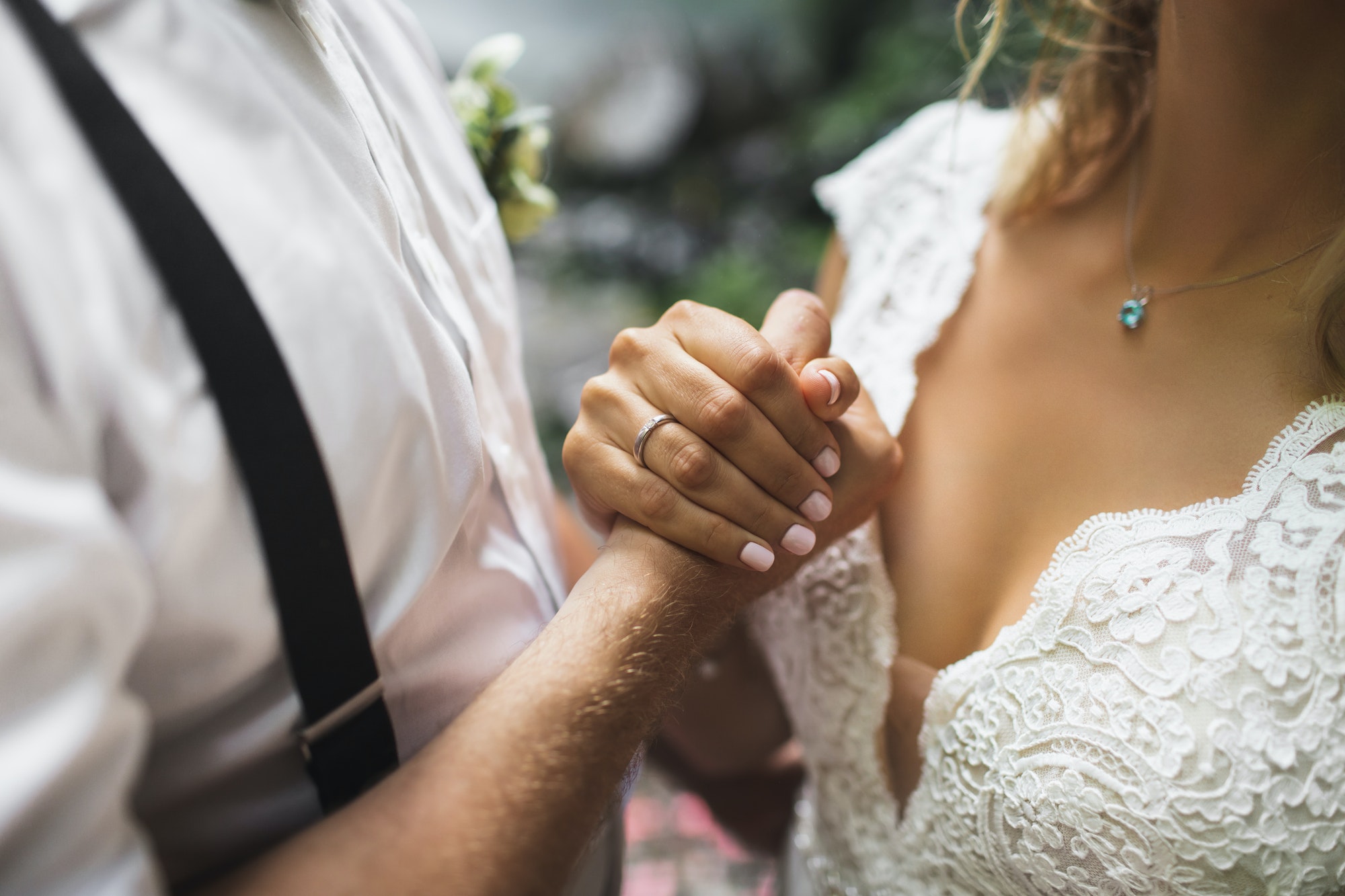 Close-up of a groom in suspenders holding the bride's hands against her lace gown