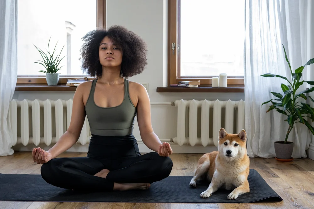 Woman seated cross-legged on a yoga mat by a window with a shiba inu beside her