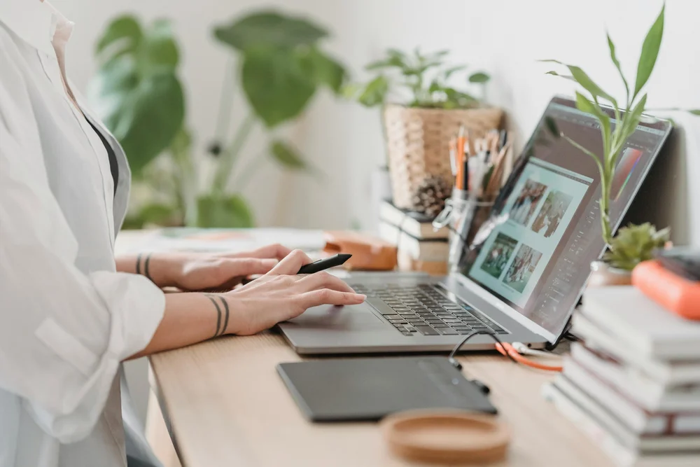 Hands at a laptop reviewing photographs on a bright desk with plants and a pen cup