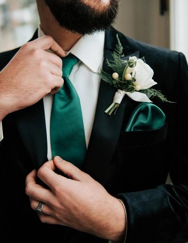 Groom adjusting an emerald green tie with a white rose boutonniere