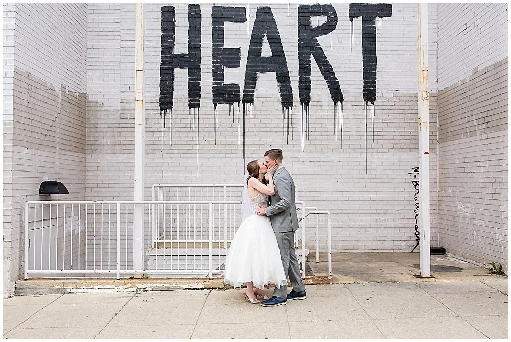 Couple embracing in front of a mural reading HEART on a white brick wall