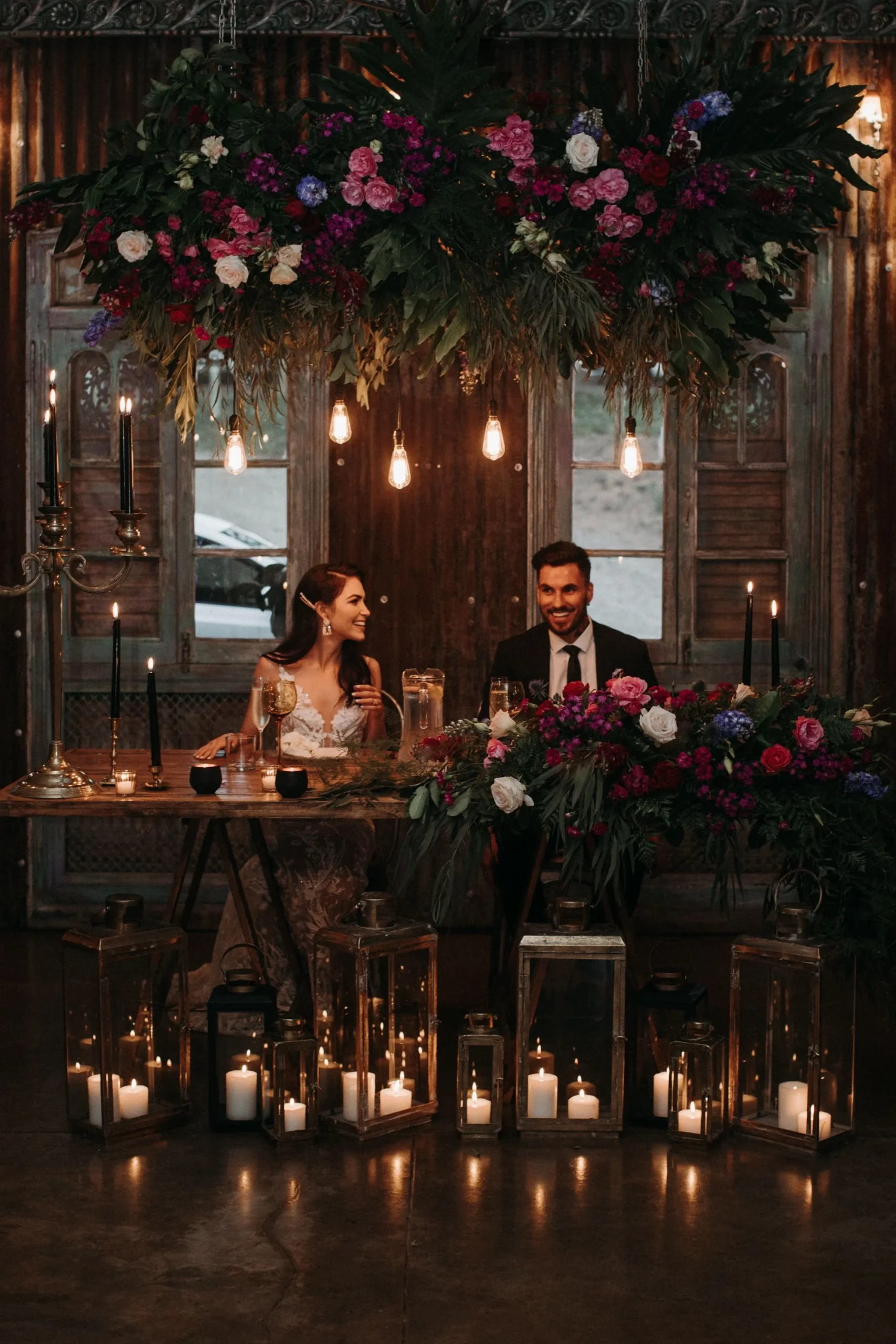 Couple seated at a candlelit table with moody florals in a rustic industrial space