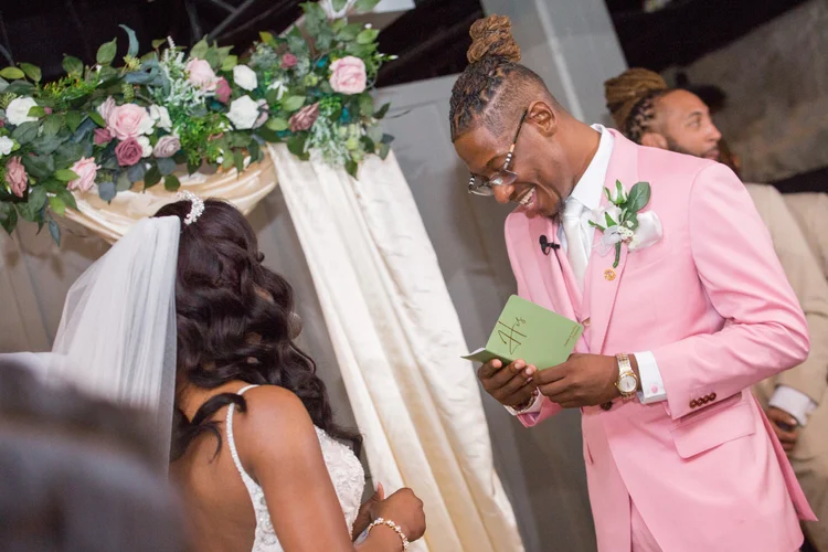 Groom in a blush suit smiling beside the floral ceremony arch