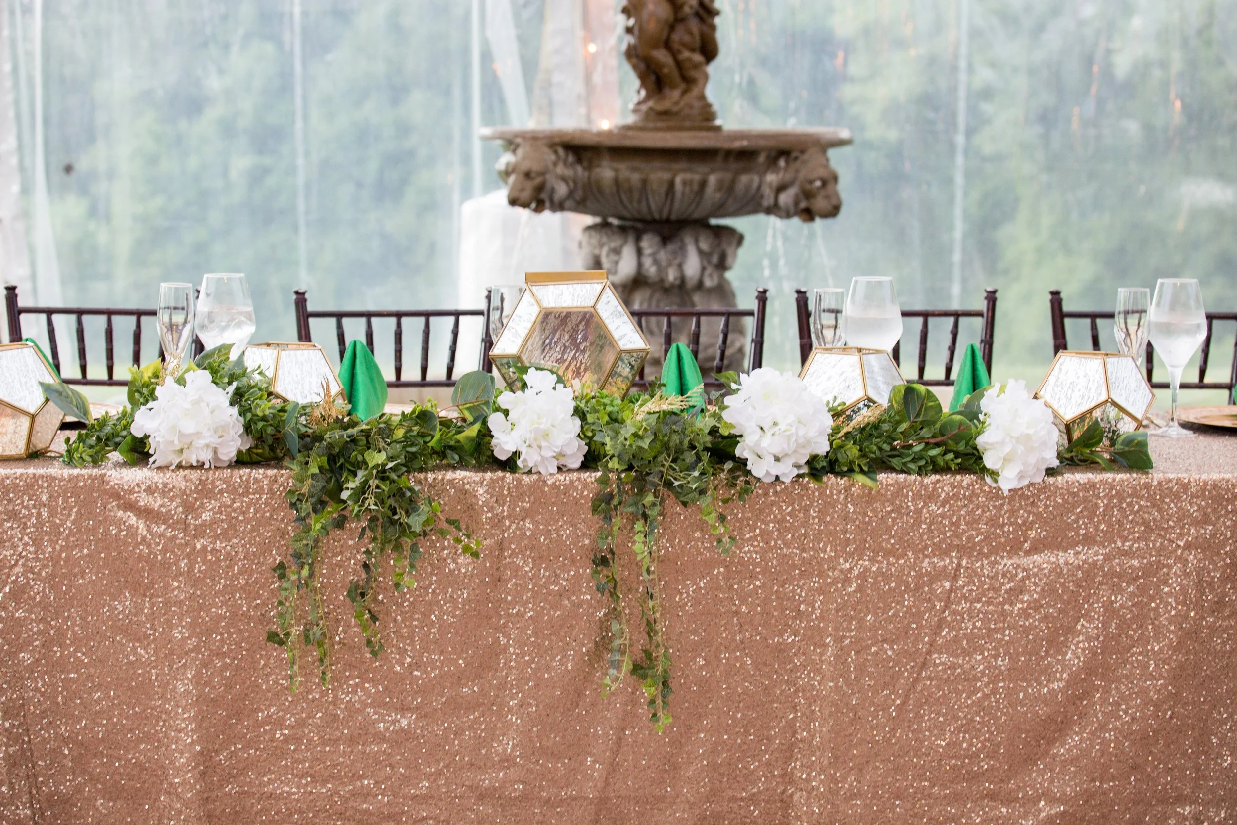 Head table with a rose gold sequin linen and white floral garland by a fountain