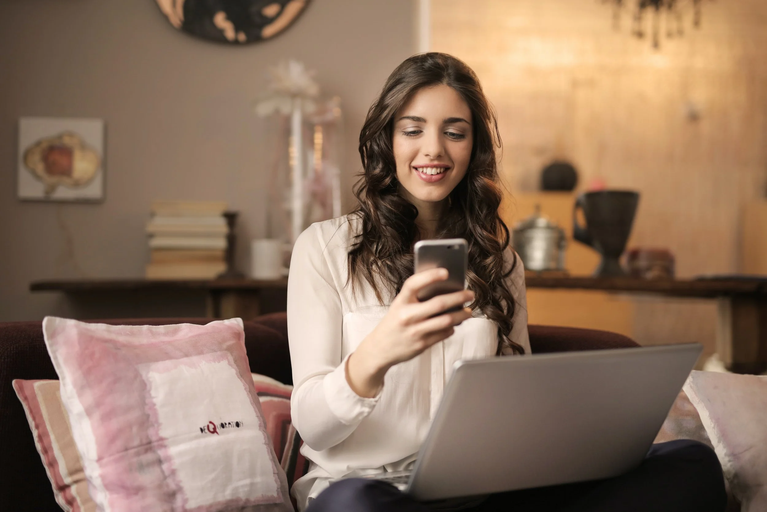Woman smiling at her phone while planning at a laptop on a sofa