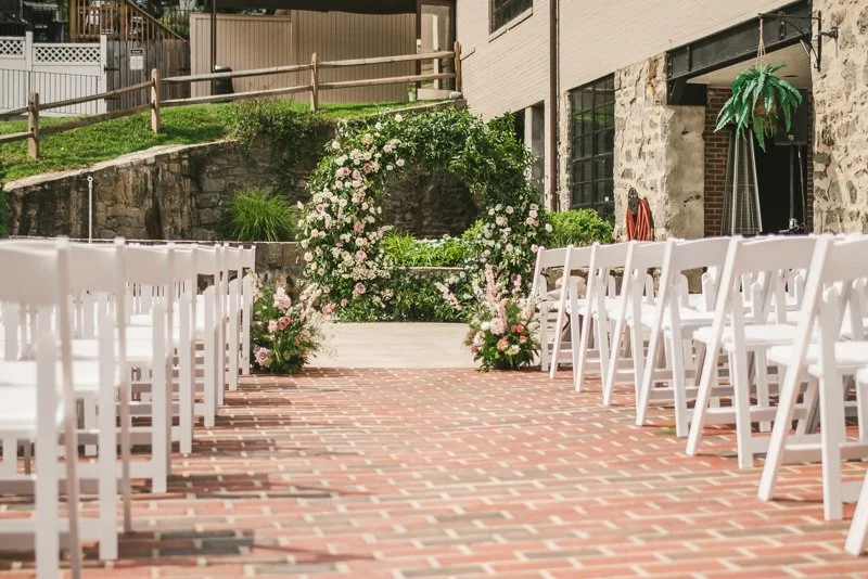 Outdoor ceremony aisle lined with white chairs and climbing roses on a brick terrace